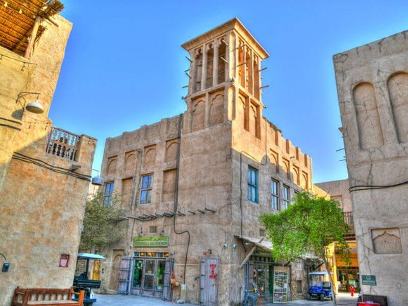A narrow street in Al Fahidi Historical Neighbourhood in Dubai, showing old sand coloured buildings with a traditional wind tower on top, small shops at street level and a green tree in front.