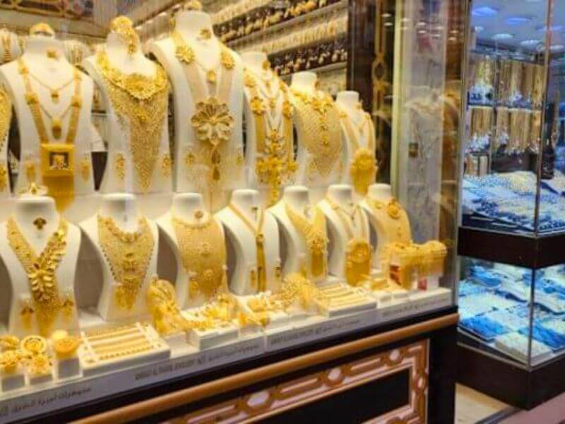 A shop window at the Gold Souk in Dubai displaying rows of gold necklaces, bracelets and jewellery sets on white mannequin stands, with more gold jewellery in glass cases on the side.