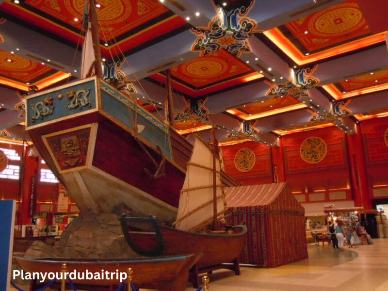 A large model of a Chinese wooden sailing ship inside the China Court section of Ibn Battuta Mall in Dubai, with a red and gold decorated ceiling above it.