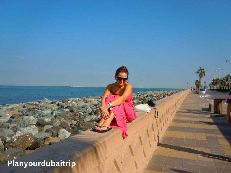 A woman in a pink dress sitting on a wall at Palm Boardwalk in Dubai, with the blue sea and rocks behind her and palm trees lining the boardwalk path.