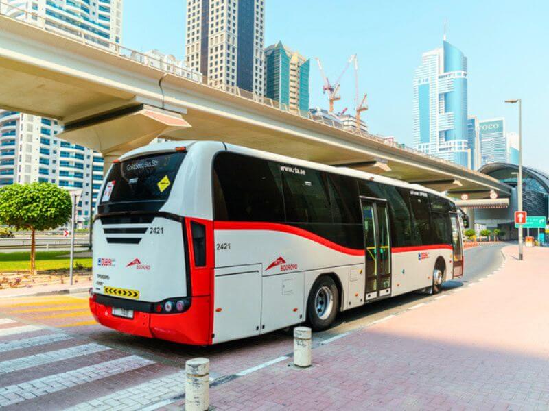 An RTA public bus stopped at a bus stop in Dubai with city skyscrapers in the background.