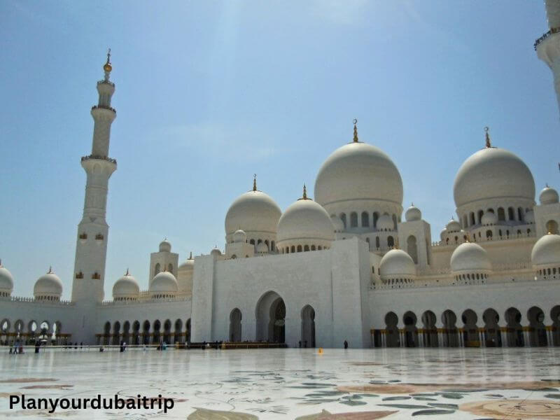 The white marble exterior of Sheikh Zayed Grand Mosque in Abu Dhabi, with its domes and minaret against a blue sky.