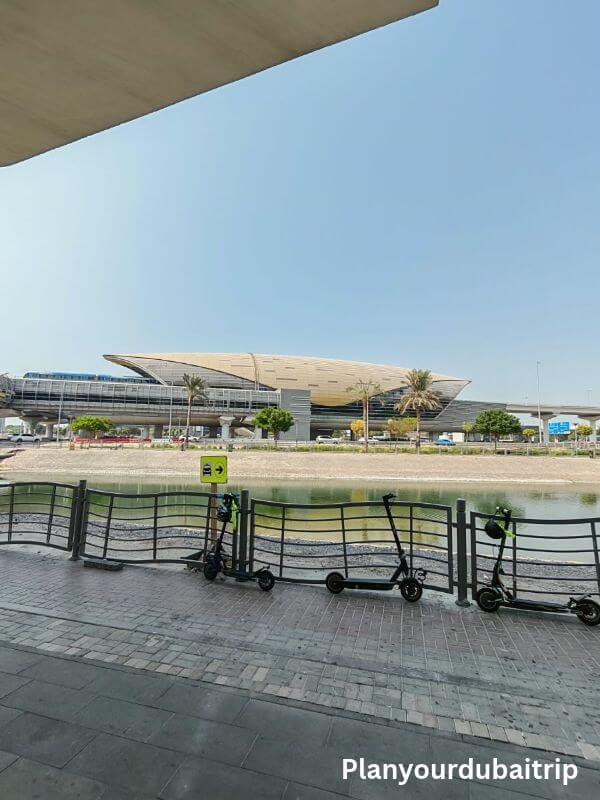 Escooters in Dubai E-scooters parked along a railing by the water in Dubai, with a metro station in the background.